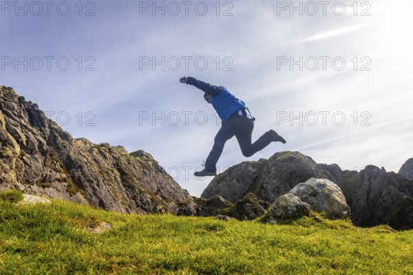 Male hiker jumping on a green meadow in front of the rocky peaks of aiako harria or penas de aia mountain in oiartzun, basque country