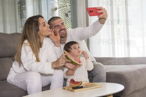 Family of three waving at camera taking a selfie while eating watermelon sitting on the sofa at home