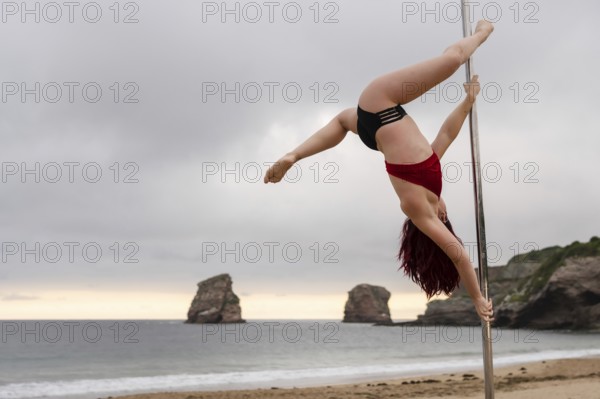 Young caucasian girl doing pole dance at sunrise at a beautiful beach. Acrobatics of pole dance face down on the sand of the beach of Hendaya, France, Europe