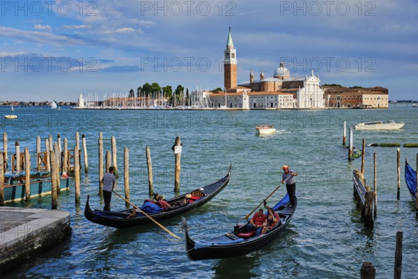 VENICE, ITALY, JUNE 27, 2018: Gondolier with tourists in gondola in lagoon of Venice by Saint Mark (San Marco) square with San Giorgio di Maggiore church in background in Venice, Italy, Europe