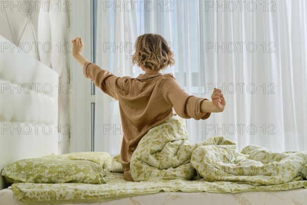 Back view of young woman stretching arms sitting on the bed at home bedroom looking to the window