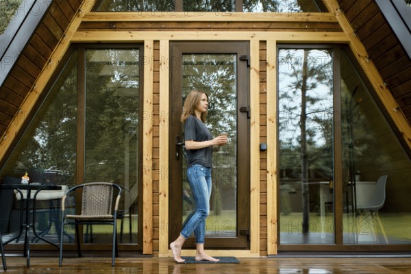 Barefoot woman came out on the wet terrace of a triangular summer house after the rain with a cup of tea in her hands