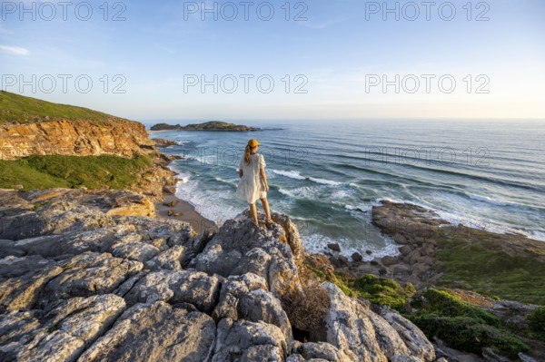 Young woman standing on a rock above the sea in the evening light, beach at The Gap, Robberg Peninsula hiking trail, Garden Route, Robberg Peninsula, Plettenberg Bay, Western Cape, South Africa, Africa