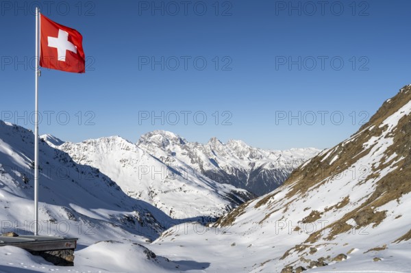 Swiss flag in front of mountain landscape in winter, mountain panorama with the peaks of the Bergüner Stöcke Tinzenhorn and Piz Ela, view at the Keschhütte in winter, Bündner Haute Route, Albula Alps, Rhaetian Alps, Grisons, Eastern Switzerland, Switzerland, Europe