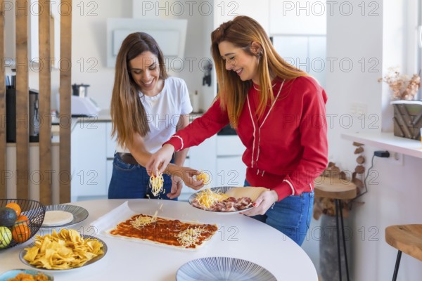 Two joyful women adding cheese to pizza dough, preparing a meal together in a modern kitchen