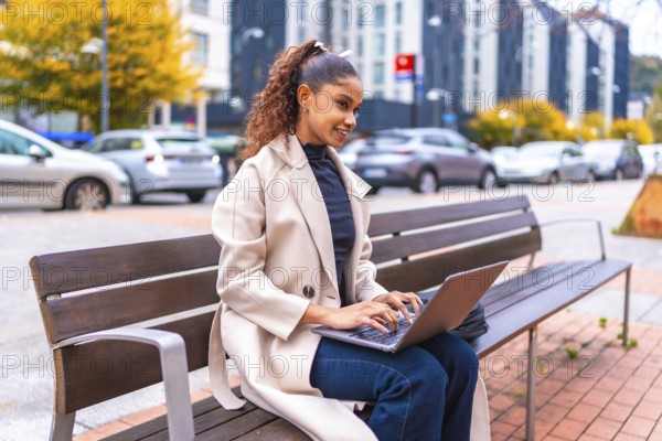 African american elegant businesswoman sitting on a bench using laptop in the city in autumn