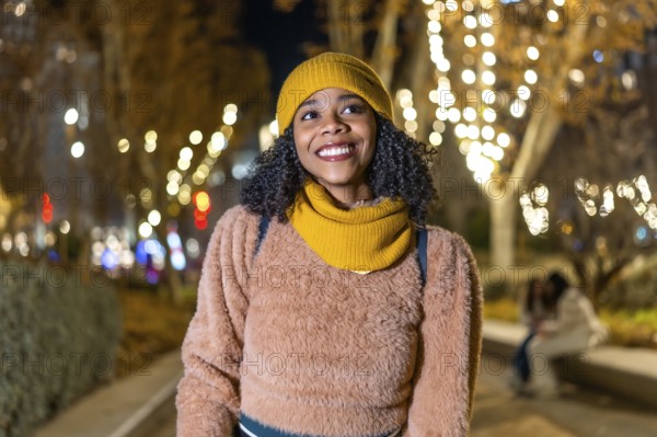 Portrait of a young black woman wearing winter clothes, smiling in a christmas decorated city at night