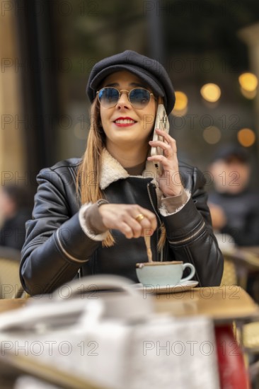 Fashionable woman wearing sunglasses and hat stirring coffee while talking on mobile phone at a parisian cafe