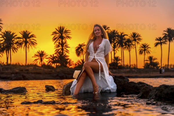Sensual blonde woman sitting on the water edge enjoying sunset in a topical beach