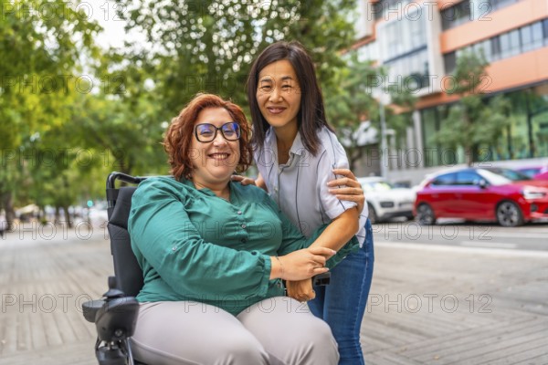 Portrait of a happy caucasian adult woman with disability sitting on electric wheelchair and her asian female caregiver smiling at camera at street