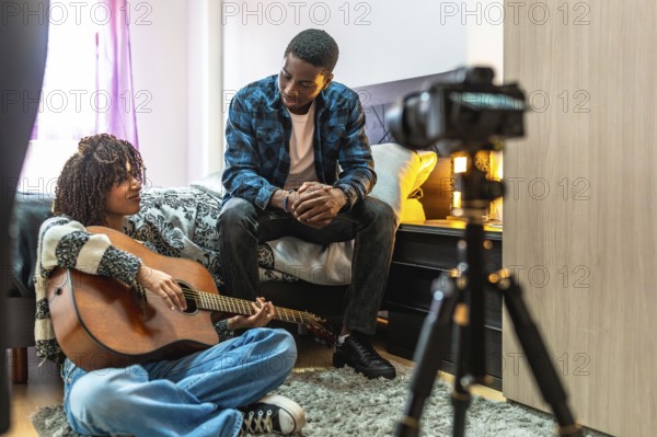 Young woman playing acoustic guitar at home with a friend composing a song and recording a video with a camera