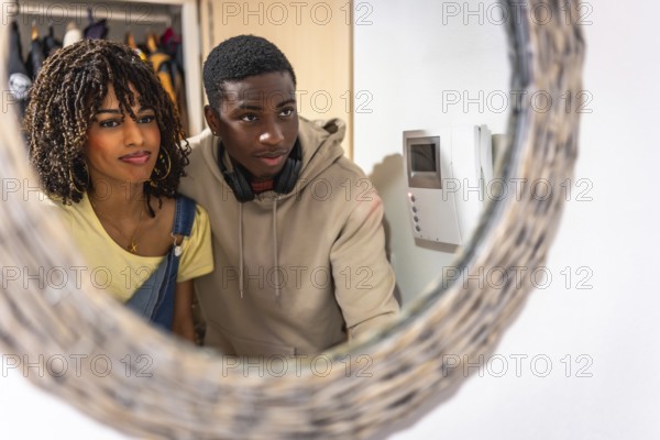 Young multiethnic couple looking at intercom screen reflected in a mirror in the entrance hall of their apartment