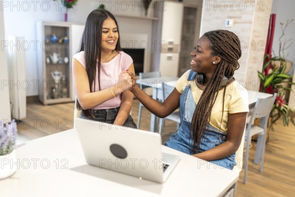 Two happy teenage girls shaking hands in agreement while using a laptop in a domestic setting, celebrating their success