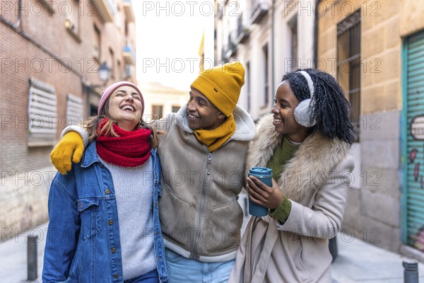 Three happy multi ethnic friends are walking embracing in the city during winter, laughing and having fun