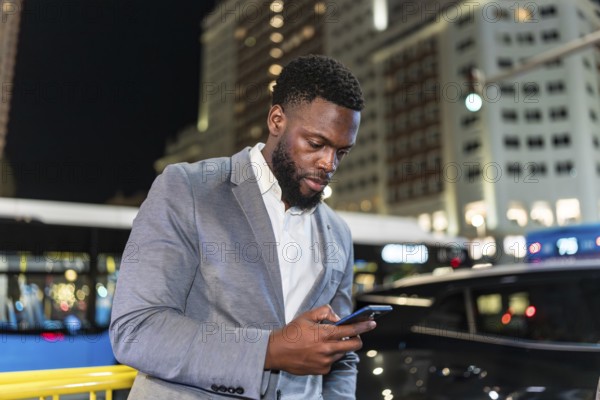 Young businessman using smartphone on a city street at night, with office buildings and traffic in the background
