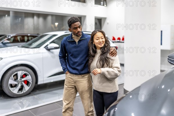 Happy multiethnic couple buying a new electric car at the dealership, embracing and choosing their future vehicle