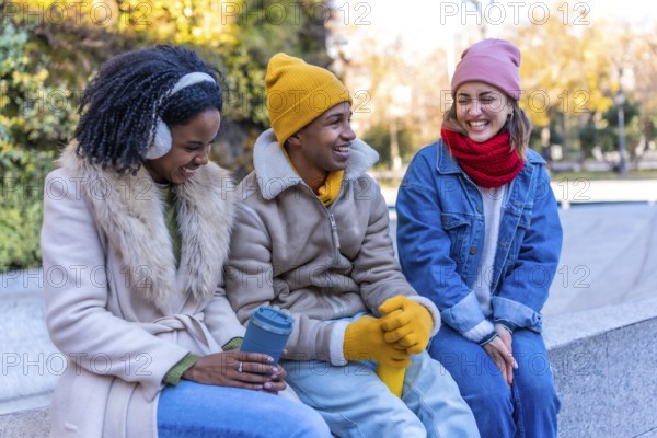 Three young multi ethnic friends wearing winter clothes chatting and laughing, sitting on a bench in a city park, during a sunny winter day