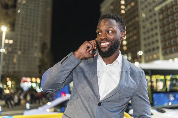 Young businessman smiling and chatting on a smartphone in a vibrant urban city at night, surrounded by dazzling lights and modern buildings