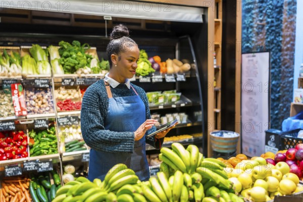 Young saleswoman using digital tablet and managing organic food in supermarket, she's wearing an apron and is standing in front of fresh fruit and vegetables