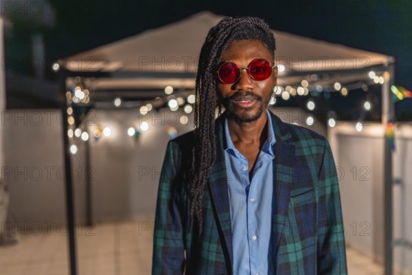 Portrait of a cool african man with afro hairslyle and colorful vintage jacket and sunglasses standing at night party in a terrace