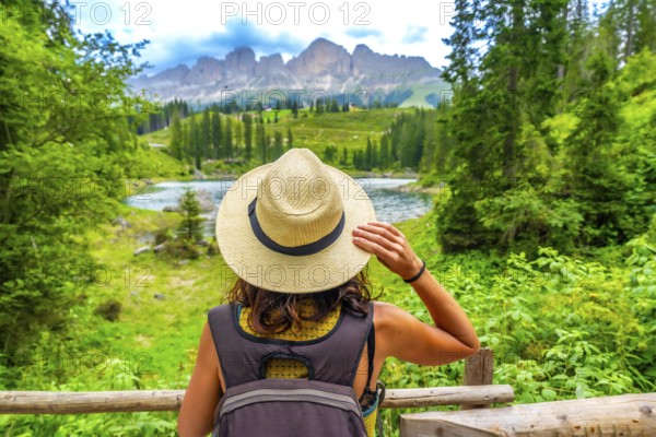 Female tourist with backpack and straw hat admiring the beautiful landscape of lake carezza and latemar mountain in the italian dolomites