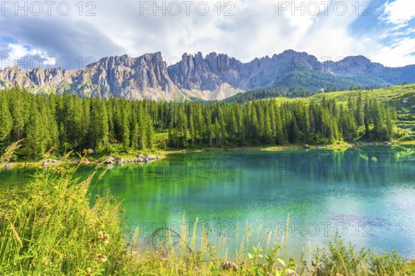 Emerald waters reflecting the latemar group and surrounding pine forest under a blue sky with clouds at karersee in summer