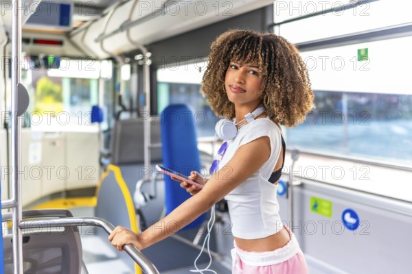 Sensual young woman with curly hair looking at camera while using phone standing in an urban bus