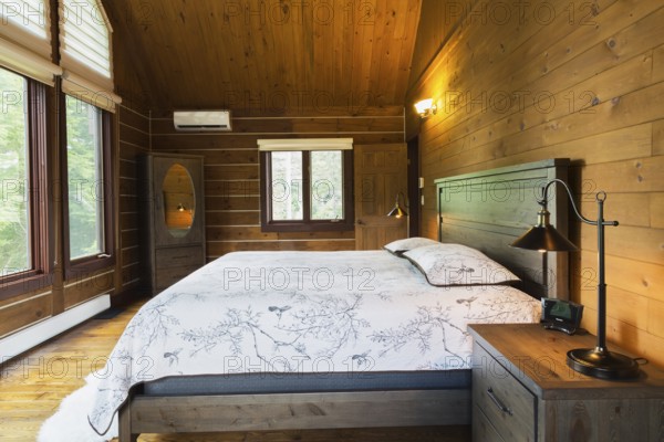 King size bed on wood-frame with wooden headboard, nightstand with antique industrial style lamp, dresser with oval mirror in master bedroom inside a milled Eastern white pine timber and flat log profile home, Quebec, Canada, North America