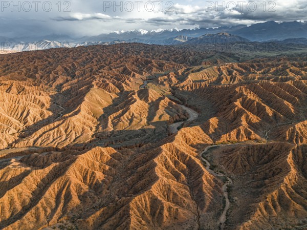 River bed runs through a landscape of eroded hills, badlands at sunset, mountain peaks of the Tian Shan Mountains in the background, aerial view, Canyon of the Forgotten Rivers, Issyk Kul, Kyrgyzstan, Asia