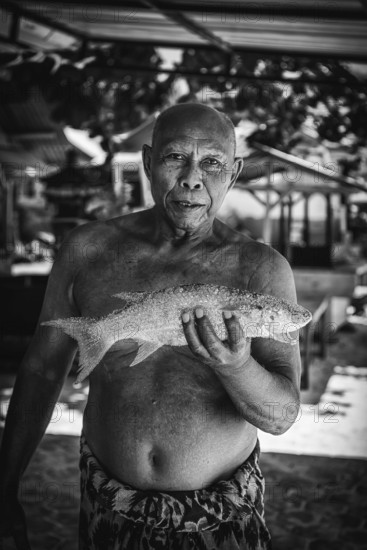 Indonesian fishmonger at a market, fish, man, food, foodstuff, local, travel, long distance, portrait, human, single, free upper body, presents, holding, showing, shows, catch, fishing, seafood, looking, joyful, market, trade, exotic, market stall, fresh, animal, sea, sea animal, black and white, monochrome, travel photo, Bali, Indonesia, Asia