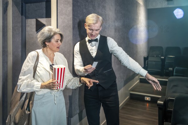 Cinema usher showing assigned seat to elegant mature woman holding popcorn bucket while entering movie theater