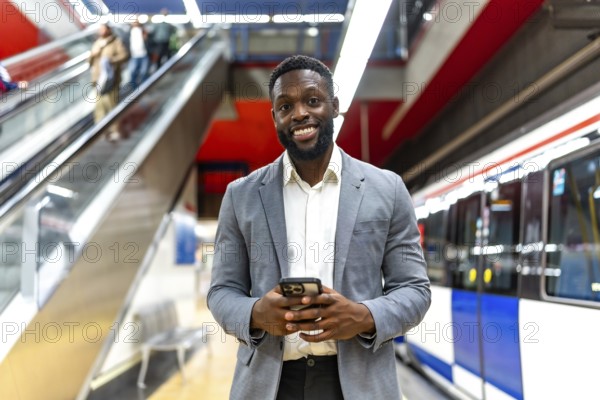 Young black businessman using smartphone while waiting for subway train in station, with escalator and train in background