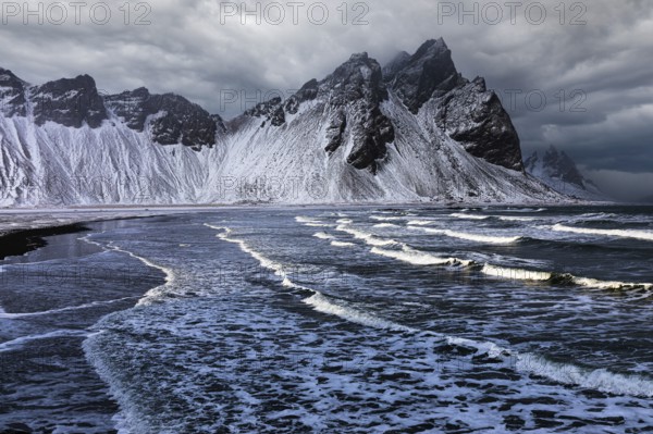White foamy waves on the beach of Stokkness, behind snowy rocky slopes of Vestrahorn, near Höfn, Sudausturland, Iceland, Europe