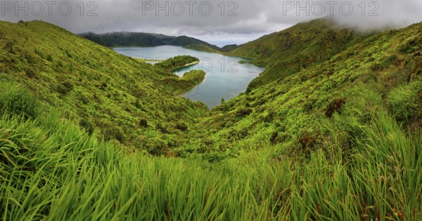 Natural landscape with a green slope and a lake surrounded by hills and a cloudy sky, crater lake Lagoa do Fogo, Caldeiras, Sao Miguel Island, Azores, Portugal, Europe