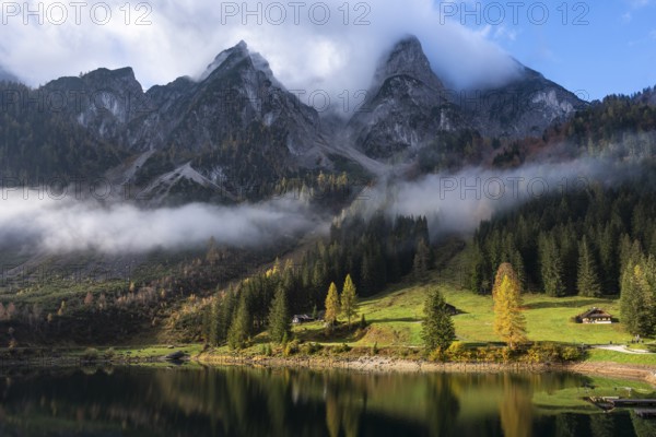 The Vordere Gosausee in autumn with a view of the cloud-covered Gosaukamm. The Seeklausalm in the centre between the trees. Cloudy and blue sky with sun. Vorderer Gosausee, Gosau, Gosau valley, Salzkammergut, Upper Austria, Austria, Europe