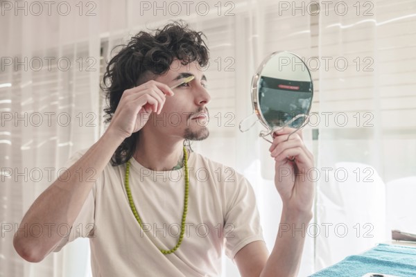 Concentrated young man with curly hair and a goatee meticulously grooming his eyebrows with tweezers while holding a small mirror in a simple home setting