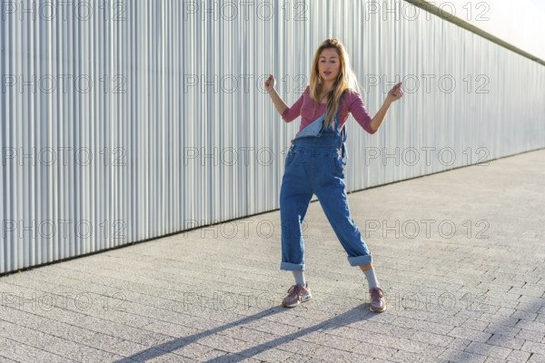 Blonde dancer performing urban dance moves on a paved area against a corrugated metal wall, showcasing energy and style