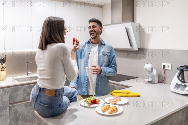 Young couple sharing a lighthearted breakfast in their contemporary kitchen, enjoying strawberries, croissants, and donuts