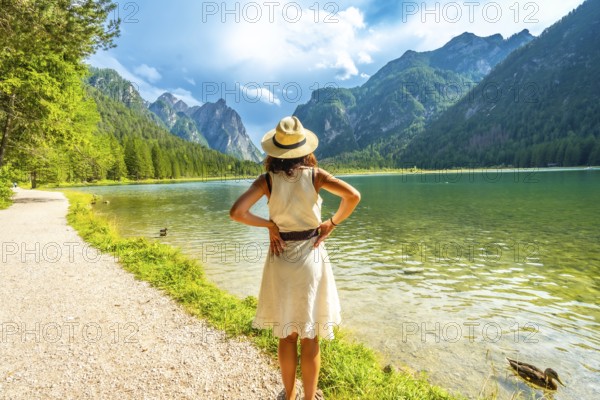 Woman wearing summer dress and straw hat admiring lake dobbiaco in the dolomites on a sunny day