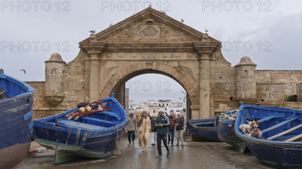 People walking through a stone arch, flanked by blue fishing boats, under a cloudy sky, Essaouira, Morocco, Africa