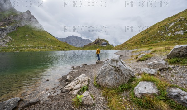 Mountaineer in yellow jacket at the mountain lake, view over the Wolayersee lake to the alpine club hut Wolayerseehütte, cloudy mountain landscape, Carnic Alps, Carnic High Trail, Carinthia, Austria, Europe