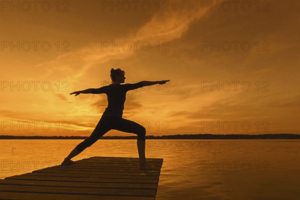 Woman practicing yoga posture Virabhadrasana II, Warrior Pose, lunging standing asana on jetty at lake at sunset
