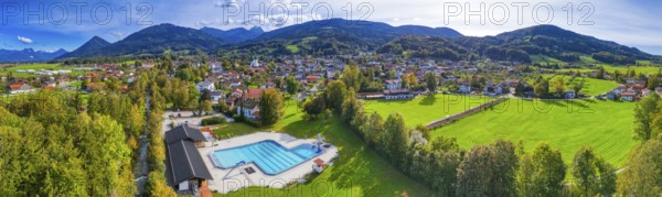 Panoramic view of a village with a swimming pool, surrounded by green meadows and forests, against a mountain backdrop, Bad Feilnbach