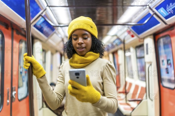 Young black woman dressed in vibrant yellow winter clothes, using her smartphone while standing inside a subway train during her commute