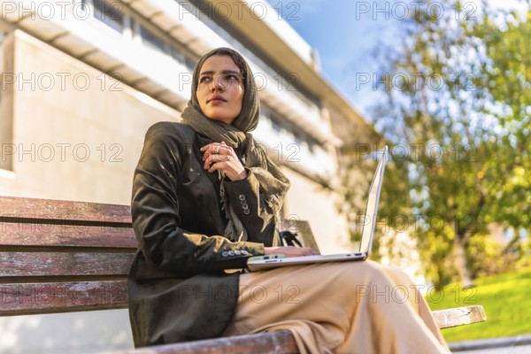 Muslim businesswoman working with laptop outdoors sitting on a bench in a park