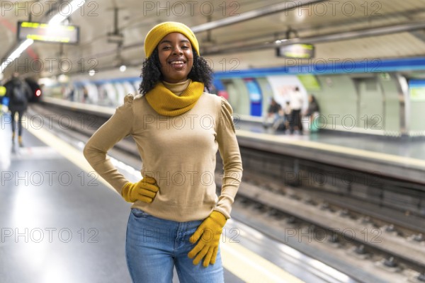 Smiling young woman wearing yellow wool cap and gloves is waiting for subway train arrival at underground station platform