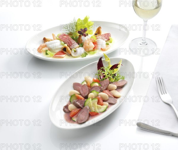 Two plates each with a meat dish and a fish dish, a glass with white wine, a fork on a napkin, white tablecloth, studio shot