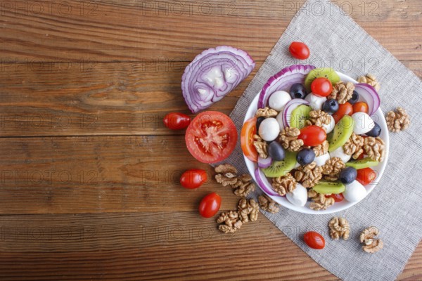 Salad with fresh cherry tomatoes, mozzarella cheese, olives, kiwi, onion and walnuts on brown wooden background, copy space, top view