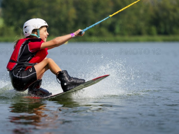 Boy or girl, sitting on wakeboard, cheerful, red life jacket, water skiing and summer holiday in wakepark, Stráž pod Ralskem, Czech Republic, Europe