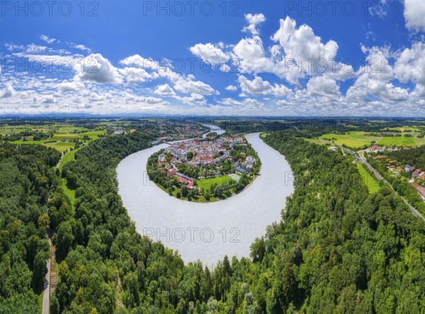 Aerial view of a village surrounded by a river, embedded in a vast green landscape under a cloudy blue sky, moated castle